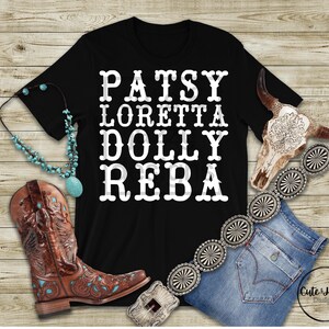 May include: Black t-shirt with white text reading "Patsy, Loretta, Dolly, Reba." The shirt is styled with a turquoise necklace, brown cowboy boots with turquoise accents, a decorative belt, and blue jeans. A decorative cow skull is also included.
