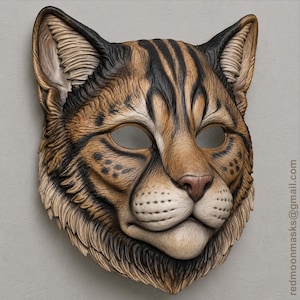 May include: A sculpted cat mask featuring brown and tan fur patterns with black stripes and spots. The mask has pointed ears, almond-shaped eye openings, and a pink nose. The text redmoonmasks@gmail.com is printed vertically on the right side.