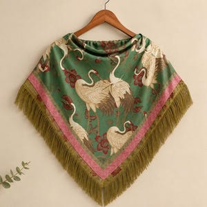 Peut inclure: Un poncho triangulaire vert orné d'un motif de grues blanches et de fleurs rouges. Le vêtement est bordé d'une bande rose et fini avec de longues franges vert olive sur les bords inférieurs, suspendu à un cintre en bois.