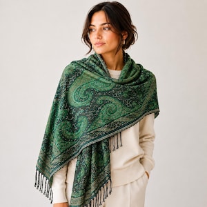 Peut inclure: Un châle rectangulaire orné d'un motif cachemire vert foncé et noir avec des volutes complexes. Le tissu présente une texture tissée et est agrémenté de franges noires sur les bords, drapé sur un haut à manches longues couleur crème.