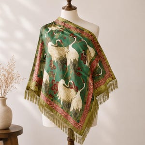 May include: A square scarf featuring a green background with a pattern of white cranes and red floral accents. The scarf has a decorative red border and is finished with long, thin green fringe along the edges, draped over a mannequin torso.