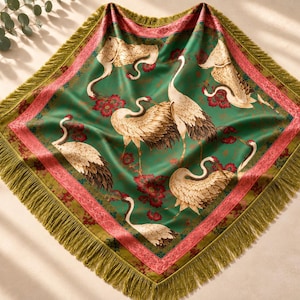May include: A square scarf featuring a vibrant green background with a repeating pattern of white cranes and red floral accents. The scarf is bordered by a pink decorative trim and finished with long, olive green fringe along all four edges.