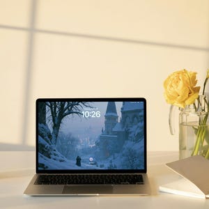 May include: A silver laptop displaying a snowy landscape with a castle on its screen, positioned next to a clear glass pitcher containing yellow flowers, a small glass candle holder, and an open white notebook on a flat surface.