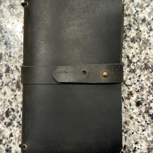 May include: A dark brown leather journal cover with a matte finish, featuring a horizontal strap closure secured by a single brass stud. The cover includes a thin black elastic cord threaded through small brass eyelets on the left side.
