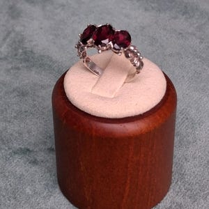 May include: A silver ring featuring three oval-cut deep red gemstones set in a row. The band has a textured, decorative metal finish along the sides, and the stones are held in place by small metal prongs, creating a classic three-stone design.