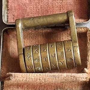 Peut inclure: Un dispositif de chiffrement en laiton composé de cinq anneaux rotatifs, chacun gravé avec diverses lettres de l'alphabet et des symboles d'étoiles, maintenus dans un cadre métallique rectangulaire. L'objet est placé dans un petit boîtier doublé.