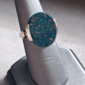 May include: A ring featuring a circular, dark green stone with small, scattered red inclusions. The stone is set in a polished silver-toned metal band that secures the gem on two sides with smooth, curved metal prongs, creating a clean, balanced look.