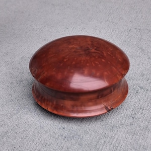 May include: A round, lidded wooden box with a polished, deep reddish-brown finish. The surface displays a natural, swirling burl wood grain pattern. The container features a smooth, curved profile with a distinct seam where the lid meets the base.