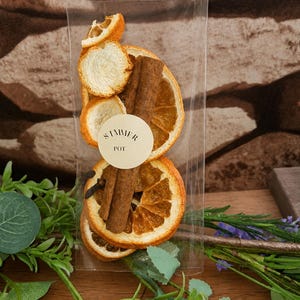 May include: A clear plastic bag containing dried orange slices, cinnamon sticks, and cloves. A round beige sticker on the front of the bag features the text SIMMER POT in black lettering. The contents are arranged vertically inside the packaging.