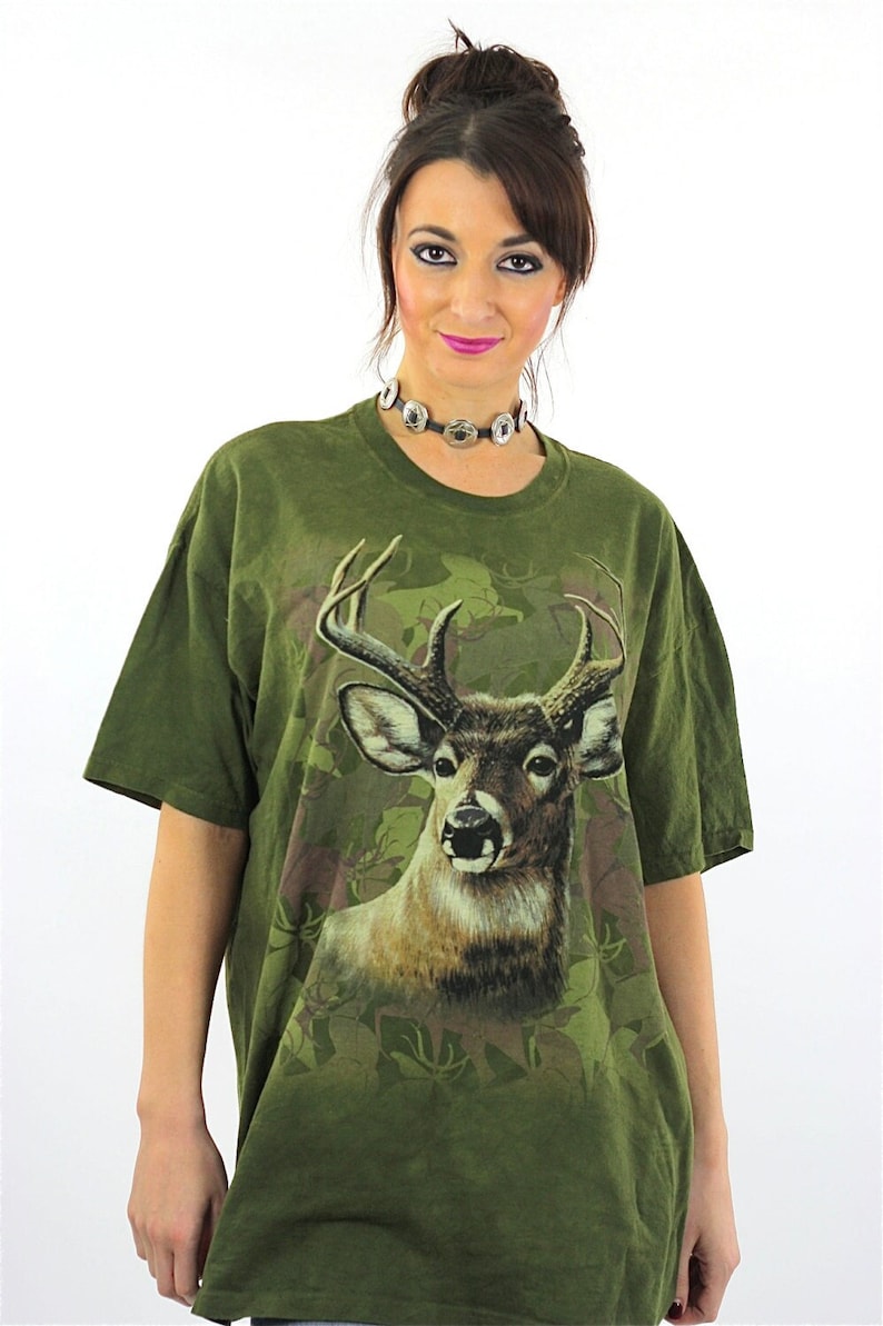 May include: A green t-shirt with a large print of a deer's head in a camouflage pattern.