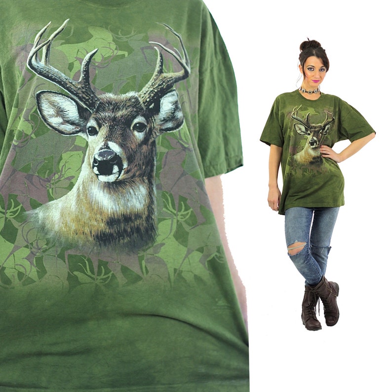 May include: Green t-shirt with a large print of a white-tailed deer in a camouflage pattern. The deer is looking directly at the viewer.