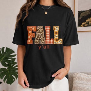 Könnte beinhalten: Ein schwarzes T-Shirt mit kurzen Ärmeln und dem Wort FALL in großen Blockbuchstaben, die jeweils ein anderes Muster aufweisen: orangefarbene Blumen, Leopardenmuster, rot-weißes Vichy-Karo und Sonnenblumen. Der Text y'all steht in Orange darunter.