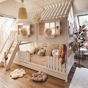 Peut inclure: Un lit superposé blanc en forme de maison avec un toit à lattes, des volets roses à découpes en forme de cœur et un escalier. Le niveau inférieur comprend un lit avec une literie beige, des barrières en palissade blanche et des peluches.