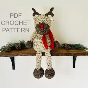 May include: A crochet reindeer doll with a beige body, dark brown hooves and muzzle, a bright red nose, and a red scarf. The text PDF CROCHET PATTERN is visible on the left side. The reindeer has antlers and is sitting upright with long legs.