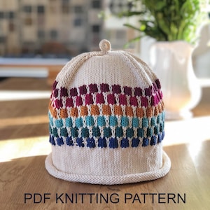 May include: A cream-colored knit hat featuring a small top knot and a multi-colored horizontal pattern of maroon, magenta, orange, light blue, teal, and dark blue stitches. The text PDF KNITTING PATTERN is printed at the bottom of the image.
