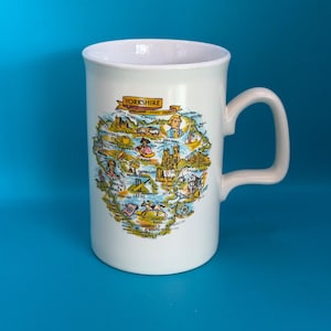 May include: A white ceramic mug featuring a colorful illustrated map of Yorkshire, England's largest county. The design includes various landmarks, figures, and the text "YORKSHIRE" and "ENGLAND'S LARGEST COUNTY" printed in a banner at the top.