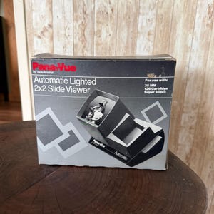 May include: A black and grey cardboard box for a Pana-Vue Automatic Lighted 2x2 Slide Viewer. The box features text stating For use with: 35 MM 126 Cartridge Super Slides and displays a graphic of the slide viewer with a black and white photo.