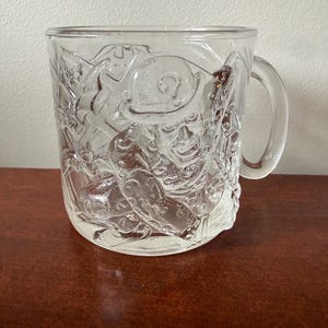 May include: A clear glass mug featuring a raised, embossed design of a character wearing a bowler hat and a suit, surrounded by question mark symbols. The mug has a smooth, curved handle on the right side and a cylindrical body with a thick base.