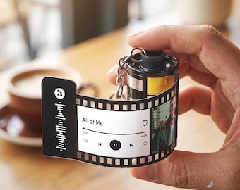 May include: A black and silver film roll keychain featuring a pull-out strip printed with a music player interface, the text All of Me, a heart icon, playback controls, a sound wave graphic, and a color photograph of a person on the end.