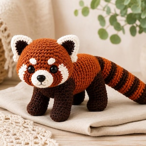 May include: A handcrafted crochet red panda plush toy featuring a rust-orange and dark brown body, white facial markings, black plastic eyes, and a long, striped tail. The stuffed animal is constructed with visible yarn stitches throughout.