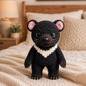 May include: A crocheted Tasmanian devil plush toy featuring black yarn, a white V-shaped chest patch, pink inner ears, dark brown snout, and black plastic eyes. The stuffed animal has white whiskers and small brown claws on its feet and hands.