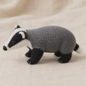 May include: A handcrafted crochet badger toy featuring a grey textured body, black legs, and a distinctive black and white striped face. The small stuffed animal has a white-tipped tail and is constructed with visible yarn stitches throughout.