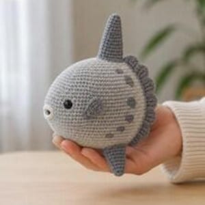 May include: A small, hand-crocheted sunfish toy made with light gray and dark gray yarn. The plush features a rounded body, a prominent dorsal fin, a small pectoral fin, and a textured tail edge, with a single black bead used for the eye.