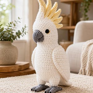 May include: A handcrafted crochet cockatoo bird toy featuring a white body, a prominent pale yellow crest on its head, a dark grey beak, and dark grey feet. The bird has black safety eyes and is constructed with visible yarn stitches throughout.