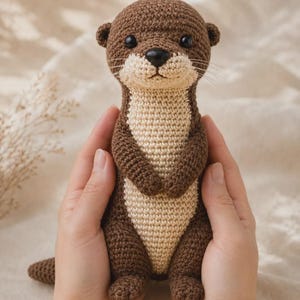 Può includere: Un giocattolo a forma di lontra realizzato all'uncinetto con filato marrone scuro per la testa, la schiena e gli arti, e filato beige chiaro per il petto e il muso. La lontra ha piccoli occhi neri in plastica, un naso nero e sottili baffi bianchi.