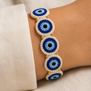 May include: A bracelet featuring five circular crocheted evil eye motifs in shades of cream, light blue, dark blue, and black. The motifs are connected by small, round, metallic gold-toned beads, creating a continuous linked design.