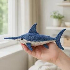 May include: A small, handcrafted crochet swordfish toy featuring a deep blue upper body and a cream-colored underbelly. The fish has a long, pointed bill, a prominent dorsal fin, a forked tail, and a small black eye, all worked in yarn.