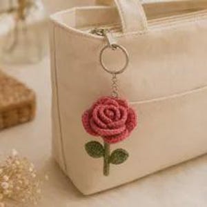 May include: A cream-colored canvas tote bag featuring a crocheted rose keychain attached to the zipper. The keychain consists of a dark pink rose blossom with a green stem and two leaves, connected to a silver-toned metal ring and chain.