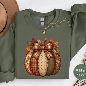 May include: A military green long-sleeve sweatshirt featuring a central graphic of a pumpkin composed of various autumn-themed fabric patterns, including plaid, floral, and polka dots, topped with a large plaid bow and colourful fall leaves.