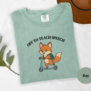 Op de afbeelding: Een bay-kleurig t-shirt met korte mouwen en een afbeelding van een cartoonvos met een rugzak op een blauwe step. Boven de vos staat de tekst OFF TO TEACH SPEECH in zwarte letters. Het shirt is voorzien van een Comfort Colors-label.