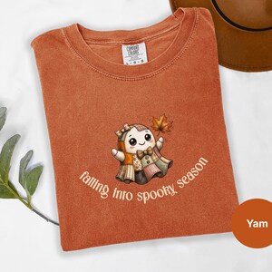 Puede incluir: Una camiseta de color óxido con un gráfico de un fantasma de patchwork que lleva una pajarita y un lazo en el pelo, sosteniendo una hoja de otoño. El texto debajo del personaje dice falling into spooky season en una fuente curva clara.