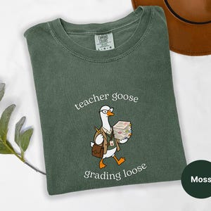 May include: A moss green crew neck t-shirt featuring a graphic of a goose wearing glasses, a tan blazer, and a lanyard, holding a stack of papers. The text above the goose reads teacher goose and the text below reads grading loose.