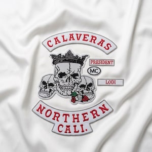 May include: A set of embroidered patches on white fabric featuring three skulls, one wearing a crown, with red roses. Text includes CALAVERAS, PRESIDENT, MC, LODI, and NORTHERN CALI. The patches are white with red and black thread detailing.