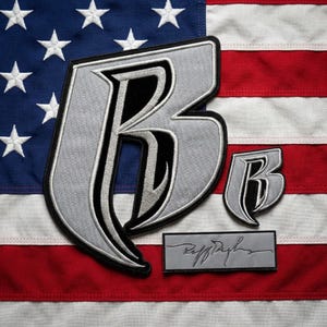 Pode incluir: Dois emblemas bordados com uma letra B estilizada em prata e preto, juntamente com um emblema retangular com uma assinatura preta sobre fundo prateado, tudo exibido sobre a bandeira americana nas cores vermelho, branco e azul.