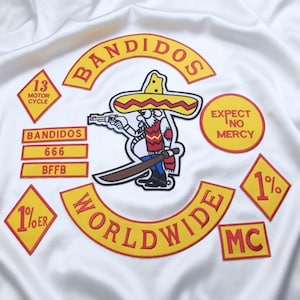 May include: A white fabric surface featuring embroidered patches with red and yellow borders. The central patch depicts a cartoon figure in a sombrero holding a pistol and machete. Text includes BANDIDOS, WORLDWIDE, 13 MOTOR CYCLE, EXPECT NO MERCY, 666, BFFB, 1%ER, 1%, and MC.
