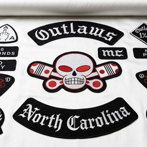 Puede incluir: Una colección de parches bordados en blanco y negro que incluye una calavera central con ojos rojos y pistones cruzados, bandas de Outlaws y North Carolina, MC, No Pity, 1%er y varios emblemas con llamas y texto.