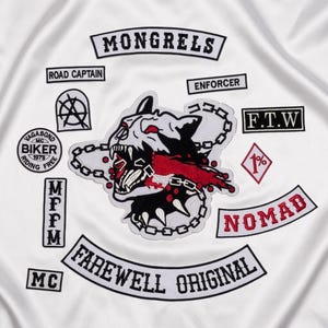 Puede incluir: Una superficie de tela blanca con varios parches bordados. Un parche central muestra una cabeza de perro gruñendo con una cadena y detalles en rojo. El texto incluye MONGRELS, ROAD CAPTAIN, ENFORCER, F.T.W, VAGABOND BIKER, MFFM, NOMAD, FAREWELL ORIGINAL y MC.