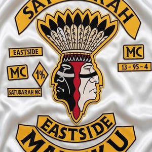 Peut inclure: Un tissu en satin blanc orné d'écussons brodés aux bordures jaunes et noires. L'emblème central représente un personnage à deux visages portant une coiffe à plumes. Le texte inclut SATUDARAH, EASTSIDE, MALUKU, MC, 1% et 13-95-4.