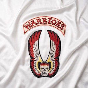 Puede incluir: Un conjunto de parches bordados que incluye una banda curva de color crema con el texto rojo WARRIORS y un emblema central de una calavera blanca con una corona de llamas rojas, flanqueada por grandes alas rojas y blancas con bordes negros.