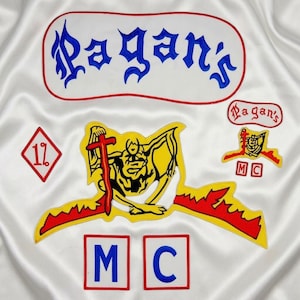 May include: A white satin fabric featuring embroidered patches including a large blue Pagan's logo, a yellow and red winged figure holding a sword, a diamond-shaped 1% patch, and two square patches with the blue letters M and C.