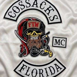 以下が含まれることがあります： 白いサテン生地に刺繍されたワッペンセット。上部のアーチにはCOSSACKS、下部のアーチにはFLORIDA、長方形のワッペンにはMCと記されています。中央のワッペンには、髭を生やしたドクロ、FTWと書かれたヘルメット、そして剣が描かれています。