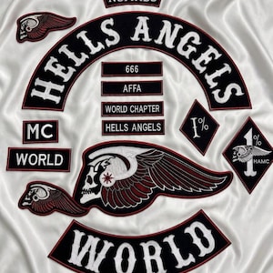 May include: A white satin fabric background featuring multiple embroidered patches. The patches include the text HELLS ANGELS, NOMADS, MC, WORLD, 666, AFFA, WORLD CHAPTER, and 1% HAMC, alongside winged skull emblems with red and white detailing.