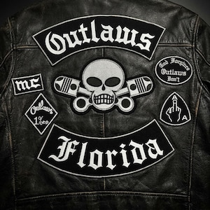 Puede incluir: Una chaqueta de cuero negro con varios parches bordados en la espalda. Los parches incluyen el texto Outlaws, Florida, MC y God Forgives Outlaws Don't, junto con un gráfico central de una calavera con pistones y un gesto de mano.