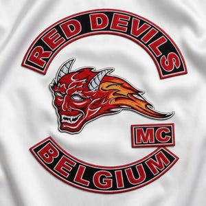 Può includere: Una superficie in tessuto bianco con toppe ricamate. Una toppa superiore arcuata riporta la scritta RED DEVILS in lettere rosse su sfondo nero. Una toppa centrale mostra una testa di diavolo rossa con fiamme e corna. In basso la scritta BELGIUM.