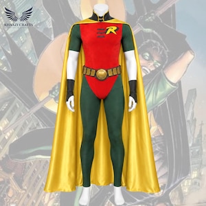 Può includere: Un costume da supereroe composto da una maglia rossa a maniche corte con un emblema R giallo, leggings verdi e un lungo mantello giallo. L'outfit include una cintura dorata con segmenti rettangolari e polsini neri su un manichino.