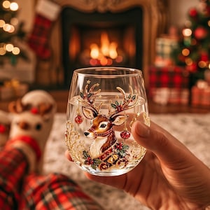 May include: A clear stemless wine glass featuring a detailed illustration of a reindeer head adorned with gold antlers, red holly berries, and hanging ornaments. The design includes intricate gold filigree accents and small golden bells.